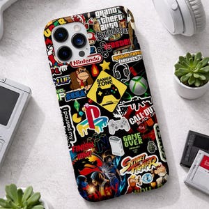 May include: Black phone case covered in colorful gaming-themed stickers, including logos for Nintendo, PlayStation, and Xbox. The case features a variety of game-related graphics and text, such as "Gamer Zone" and "Grand Theft Auto".