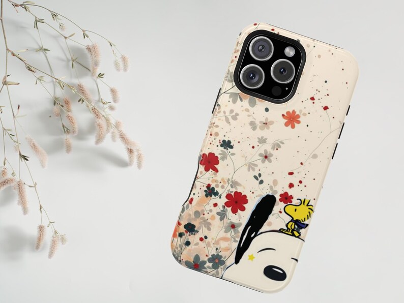 May include: A phone case featuring a floral pattern and a cartoon illustration of Snoopy and Woodstock. The case has a cream-coloured background with red, grey, and white flowers. Snoopy and Woodstock are on the bottom right.