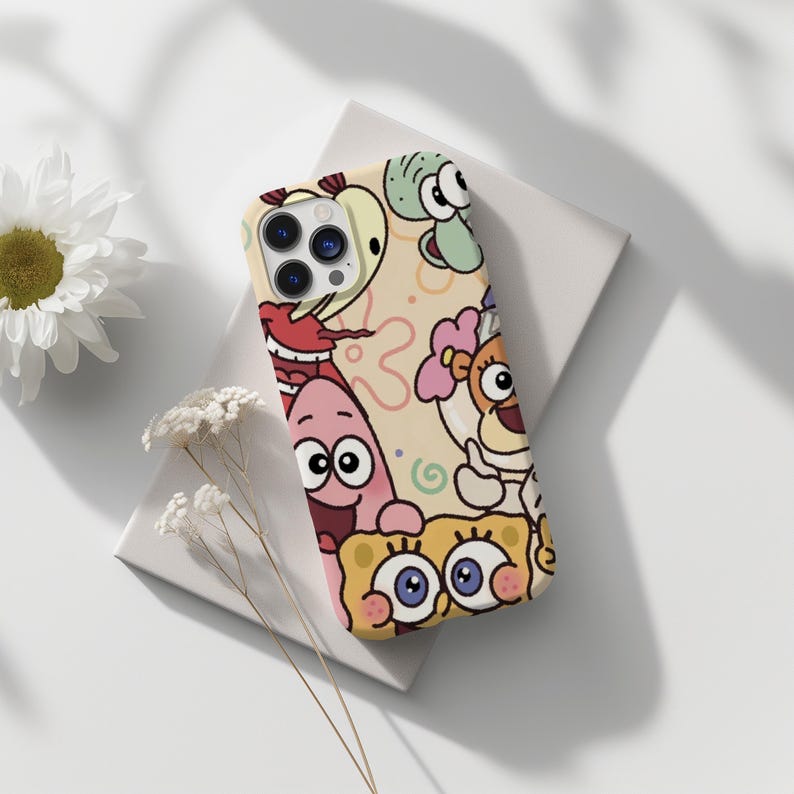 May include: A phone case featuring cartoon characters on a light tan background. The case has a colorful design with characters like SpongeBob SquarePants, Patrick Star, and Squidward Tentacles. The case is on a white surface.