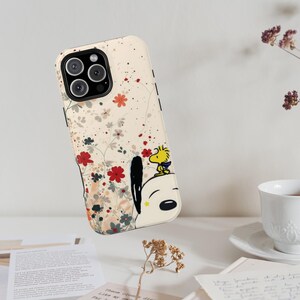 May include: A cream-coloured smartphone case featuring a graphic of Snoopy and Woodstock surrounded by delicate red, grey, and orange floral illustrations. The case has a black camera cutout and a smooth, matte-finish surface design.