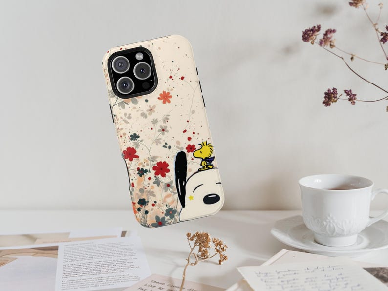 May include: A cream-coloured smartphone case featuring a graphic of Snoopy and Woodstock surrounded by delicate red, grey, and orange floral illustrations. The case has a black camera cutout and a smooth, matte-finish surface design.