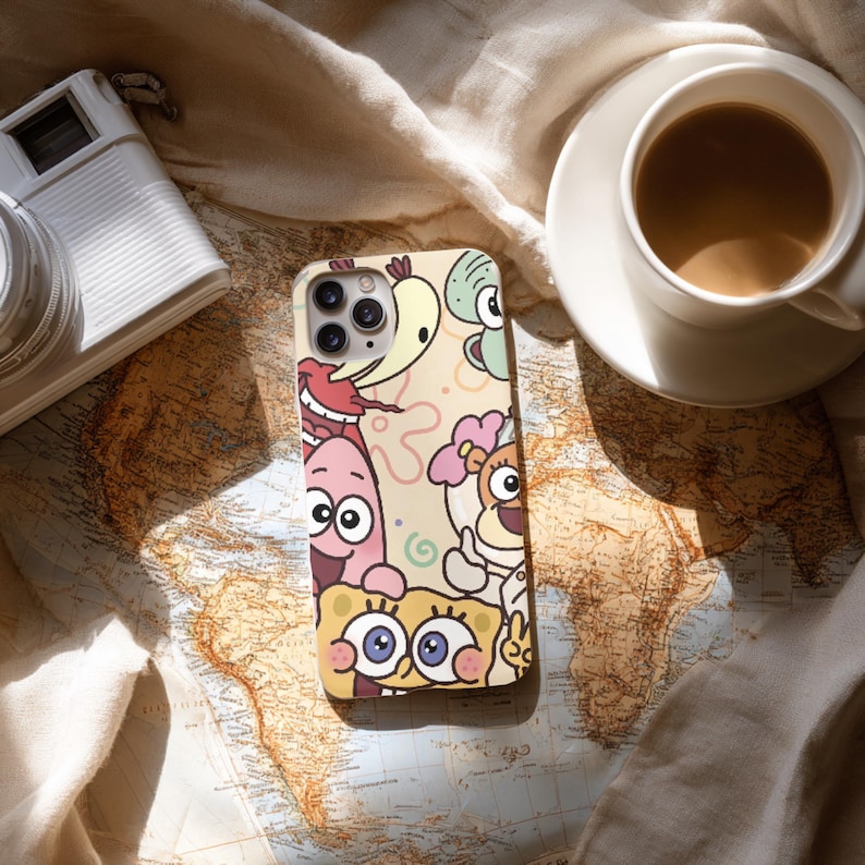 May include: A beige phone case featuring cartoon characters, including SpongeBob SquarePants and Patrick Star. The case is on a world map, next to a camera and a cup of coffee.