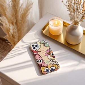 May include: A phone case featuring a colorful cartoon design with various characters. The case has a cream-colored background with images of cartoon characters in shades of yellow, pink, blue, and brown. The phone case is on a white surface.