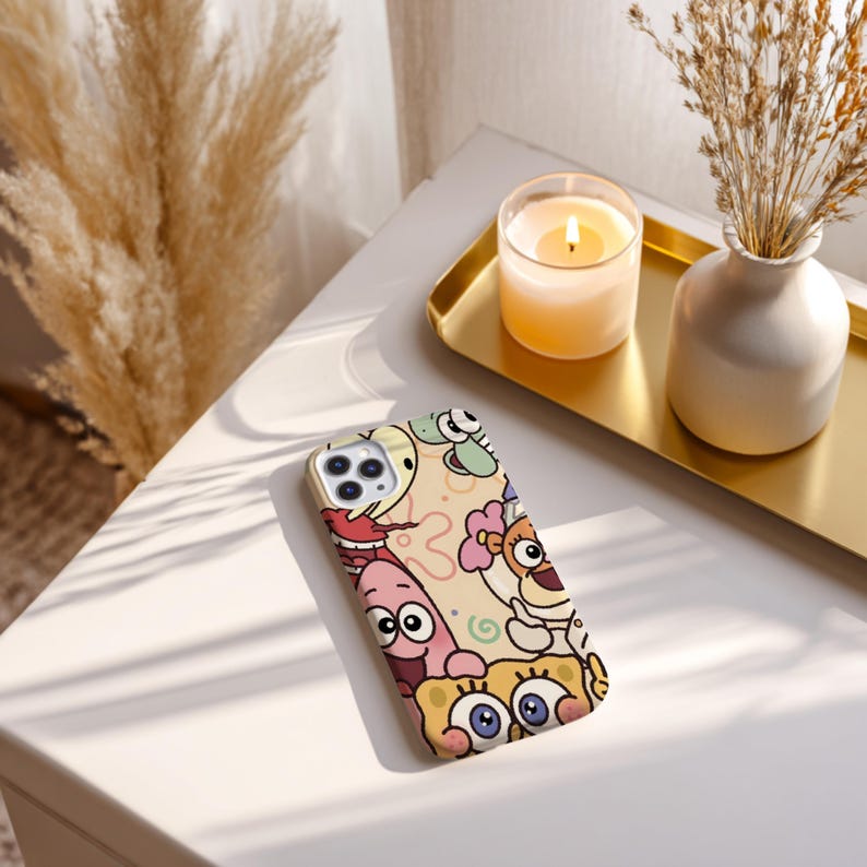 May include: A phone case featuring a colorful cartoon design with various characters. The case has a cream-colored background with images of cartoon characters in shades of yellow, pink, blue, and brown. The phone case is on a white surface.
