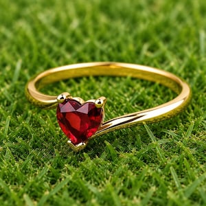 May include: A gold ring featuring a single heart-shaped red gemstone set in a polished metal band. The ring has a smooth, reflective finish and a delicate, slender profile, showcasing the vibrant crimson color of the faceted heart stone.