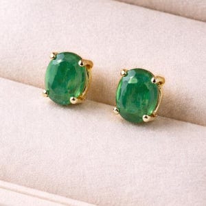 May include: A pair of oval-shaped green gemstone stud earrings set in yellow gold prongs. The earrings are displayed side-by-side, each featuring a faceted green stone secured by four small gold prongs, showcasing a polished and reflective surface.