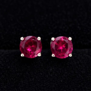 May include: A pair of round, vibrant red gemstone stud earrings. The gemstones are set in a simple, four-prong setting against a dark background. The earrings are a classic jewelry item.
