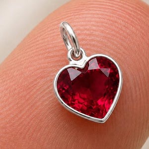 May include: A small heart-shaped pendant featuring a deep red faceted gemstone set in a polished silver-toned metal frame. The charm includes a small circular jump ring at the top for attachment, showcasing a clear, vibrant red colour and precise cut.