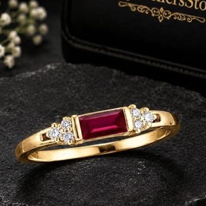 Può includere: Un anello d'oro con una gemma rettangolare rosso scuro al centro, affiancata da gemme più piccole e chiare. L'anello è posto su una superficie scura e strutturata. Il nome del marchio "RajjewellersStore" è visibile in lettere dorate.