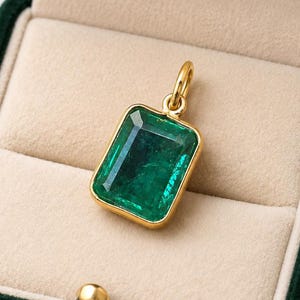 May include: A rectangular emerald pendant with a gold setting is displayed in a dark green velvet jewellery box. The pendant's emerald is a vibrant green colour. The box has a beige interior and a gold clasp.
