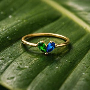May include: A gold ring featuring a green and blue gemstone design. The ring is set against a green leaf background.