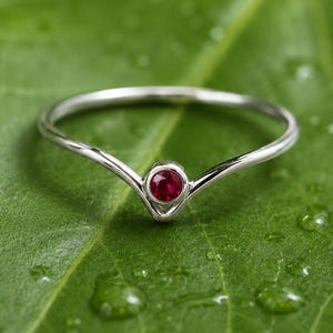 Könnte beinhalten: Ein silberfarbener V-förmiger Ring mit einem einzelnen, kleinen, runden, leuchtend roten Edelstein in der Mitte. Das Band ist dünn und glatt, wobei das Metall eine Chevron- oder V-Form bildet, die den kreisförmigen roten Stein sicher hält.