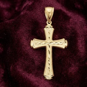 May include: A gold cross pendant featuring a textured surface with diamond-cut detailing along the centre bars. The pendant includes a polished tapered bail at the top for attachment to a chain, showcasing a bright metallic finish throughout.