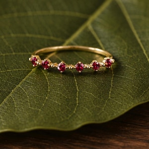 May include: A delicate gold ring featuring a row of small, vibrant red gemstones. The ring is set against a green leaf and a wooden surface, creating a natural and elegant aesthetic. The gemstones are evenly spaced along the band.