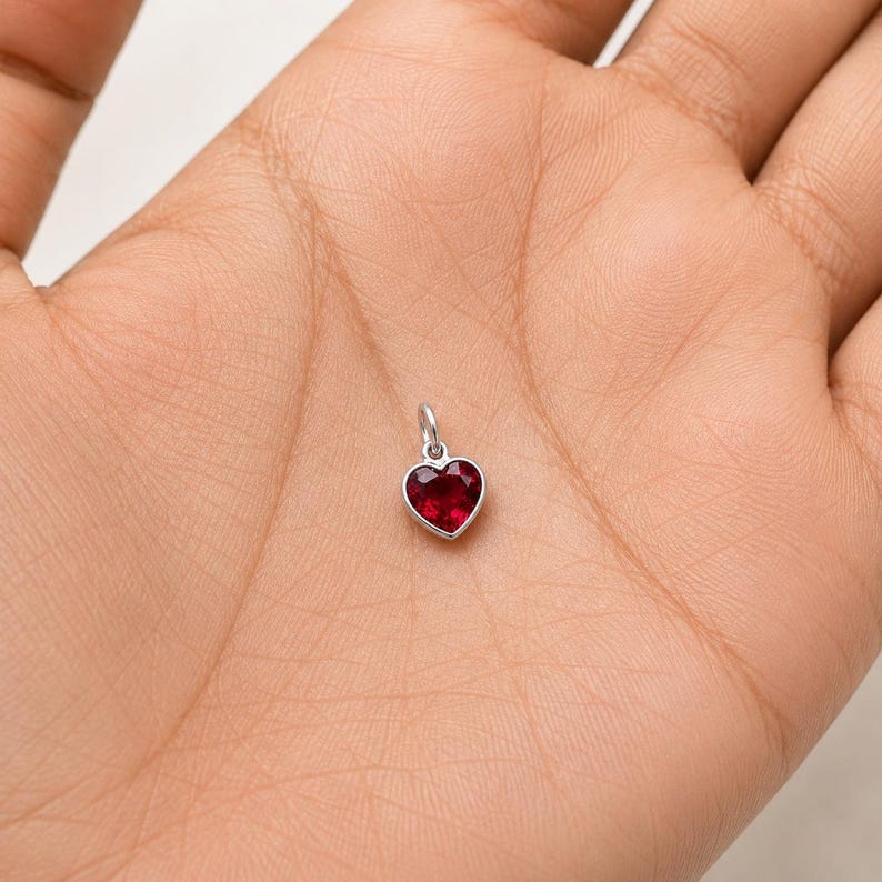May include: A small heart-shaped pendant featuring a faceted deep red gemstone set in a polished silver-toned metal frame. The charm includes a small circular jump ring at the top for attachment, showcasing a clear, vibrant red colour and smooth edges.
