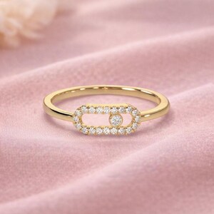 Puede incluir: Un anillo de oro con un dise&ntilde;o ovalado central incrustado con peque&ntilde;os diamantes brillantes. Un solo diamante, m&aacute;s grande, est&aacute; engastado en el centro del &oacute;valo. La banda del anillo es de oro pulido. El anillo se muestra sobre una superficie rosa.