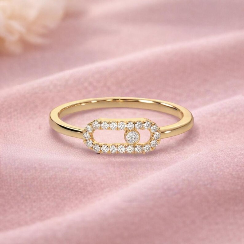 Puede incluir: Un anillo de oro con un dise&ntilde;o ovalado central incrustado con peque&ntilde;os diamantes brillantes. Un solo diamante, m&aacute;s grande, est&aacute; engastado en el centro del &oacute;valo. La banda del anillo es de oro pulido. El anillo se muestra sobre una superficie rosa.