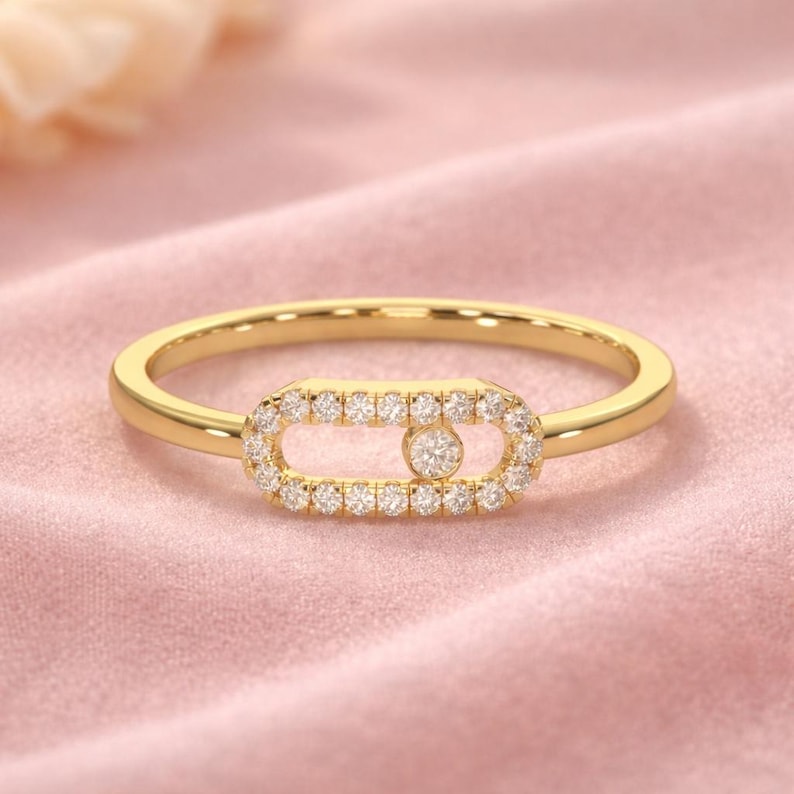 Puede incluir: Un anillo de oro con un dise&ntilde;o central rectangular. El centro est&aacute; delineado con peque&ntilde;as piedras claras, y una piedra m&aacute;s grande se encuentra en el medio. El anillo est&aacute; sobre un fondo rosa.
