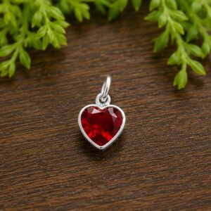 May include: A heart-shaped pendant featuring a faceted deep red gemstone set in a polished silver-toned metal frame. The pendant includes a small circular jump ring at the top for attachment, showcasing a clear, brilliant cut and a smooth, reflective surface.