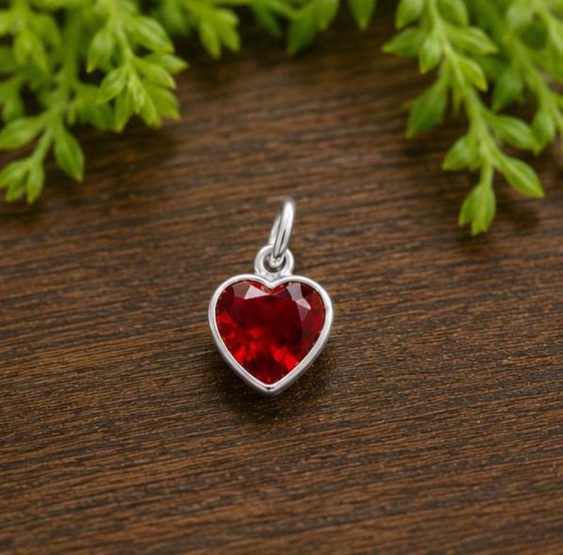 May include: A heart-shaped pendant featuring a faceted deep red gemstone set in a polished silver-toned metal frame. The pendant includes a small circular jump ring at the top for attachment, showcasing a clear, brilliant cut and a smooth, reflective surface.