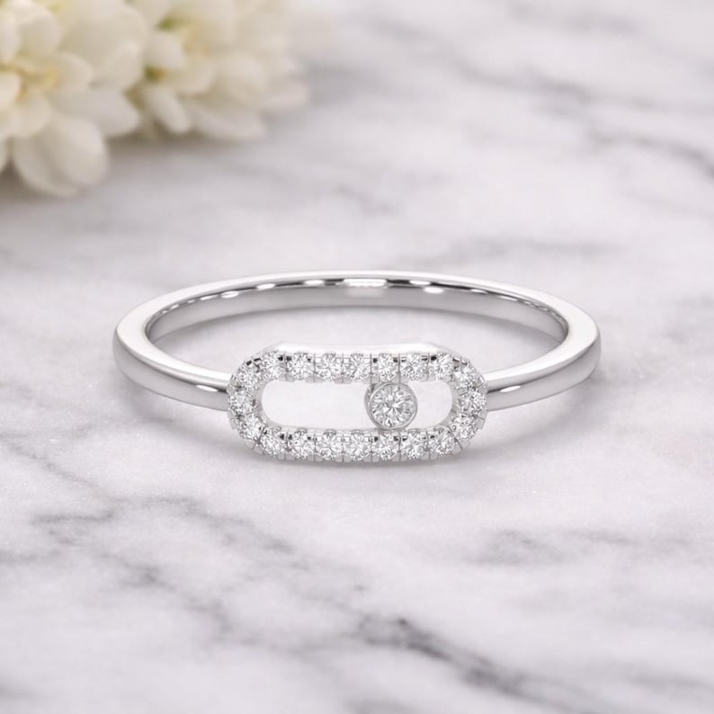 Puede incluir: Anillo de plata con un centro rectangular engastado con diamantes. Un solo diamante est&aacute; engastado en el centro del rect&aacute;ngulo. El anillo est&aacute; sobre un fondo de m&aacute;rmol blanco, con flores blancas en el fondo.