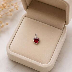 May include: A small heart-shaped pendant featuring a faceted deep red gemstone set in a polished silver-toned metal frame with a circular jump ring at the top, presented inside a beige velvet-lined jewellery box with a hinged lid.