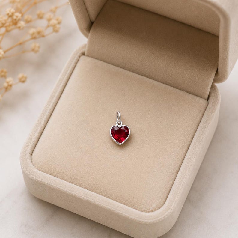 May include: A small heart-shaped pendant featuring a faceted deep red gemstone set in a polished silver-toned metal frame with a circular jump ring at the top, presented inside a beige velvet-lined jewellery box with a hinged lid.