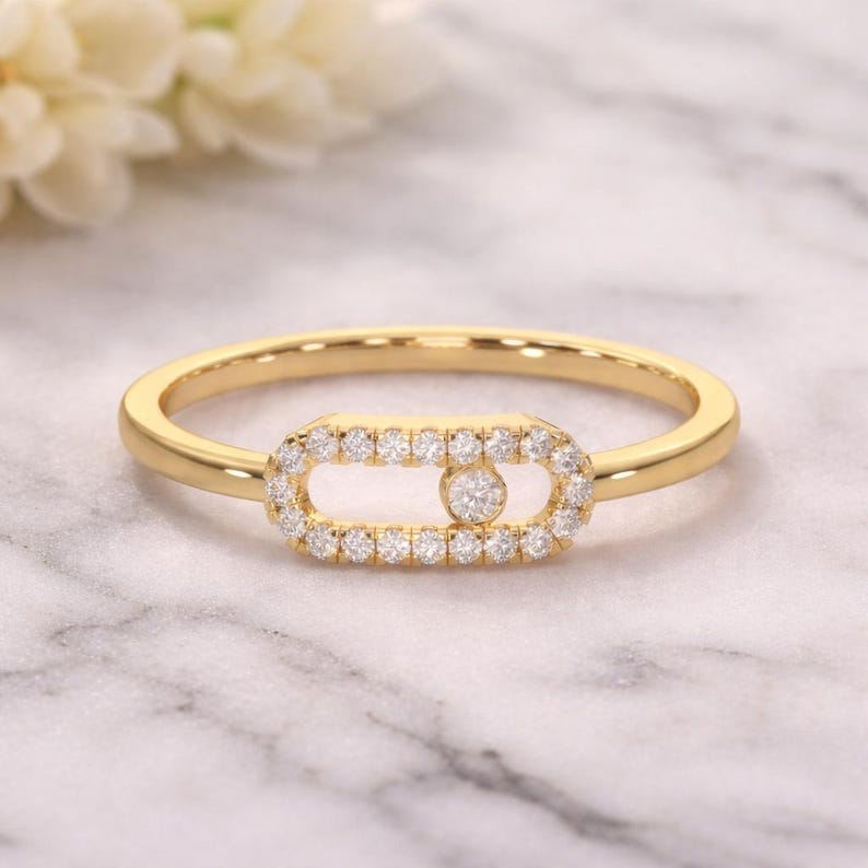 Puede incluir: Un anillo de oro con un centro rectangular, incrustado con peque&ntilde;as piedras preciosas transparentes. Una sola piedra preciosa m&aacute;s grande se encuentra en el centro del rect&aacute;ngulo. La banda del anillo es de oro pulido. El anillo est&aacute; sobre un fondo de m&aacute;rmol.