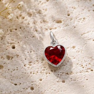 May include: A silver heart-shaped pendant featuring a faceted deep red gemstone centre. The pendant includes a small silver jump ring at the top for attachment to a chain. The design is simple, focusing on the contrast between the metallic silver setting and the vibrant red stone.