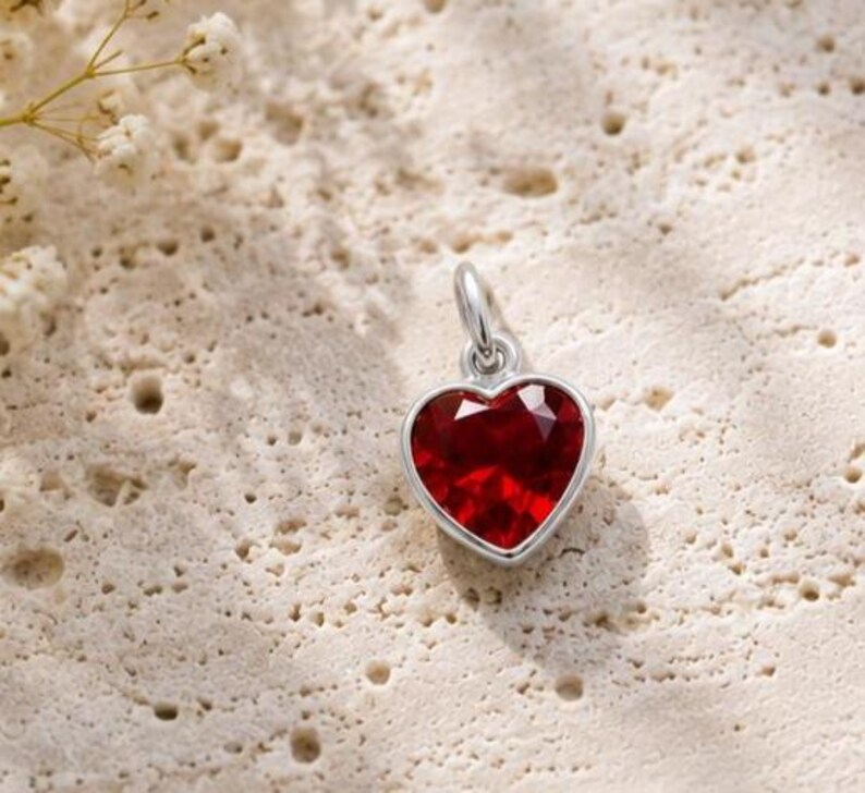 May include: A silver heart-shaped pendant featuring a faceted deep red gemstone centre. The pendant includes a small silver jump ring at the top for attachment to a chain. The design is simple, focusing on the contrast between the metallic silver setting and the vibrant red stone.