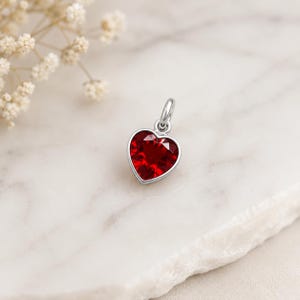 May include: A silver-toned pendant featuring a faceted, deep red heart-shaped gemstone set within a polished metal frame. The pendant includes a small circular jump ring at the top for attachment, showcasing a clear, vibrant crimson colour.