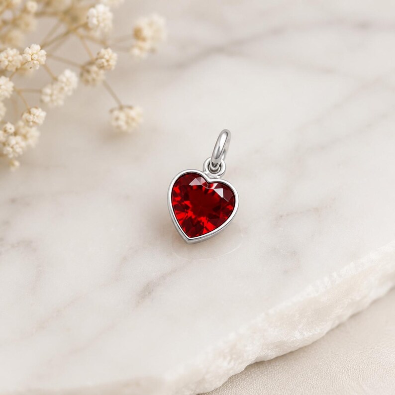 May include: A silver-toned pendant featuring a faceted, deep red heart-shaped gemstone set within a polished metal frame. The pendant includes a small circular jump ring at the top for attachment, showcasing a clear, vibrant crimson colour.