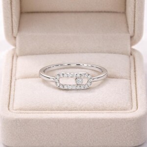 Puede incluir: Un anillo de plata con un centro rectangular con incrustaciones de diamantes. El centro presenta un solo diamante m&aacute;s grande en el medio. El anillo est&aacute; colocado sobre una caja de joyer&iacute;a beige suave.