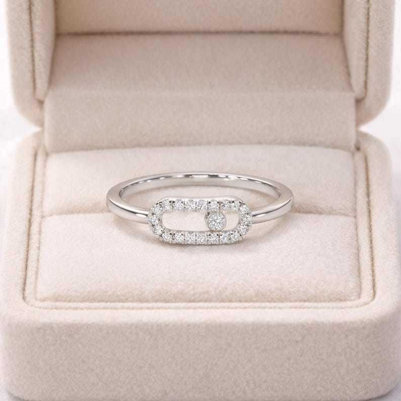 Puede incluir: Un anillo de plata con un centro rectangular con incrustaciones de diamantes. El centro presenta un solo diamante m&aacute;s grande en el medio. El anillo est&aacute; colocado sobre una caja de joyer&iacute;a beige suave.