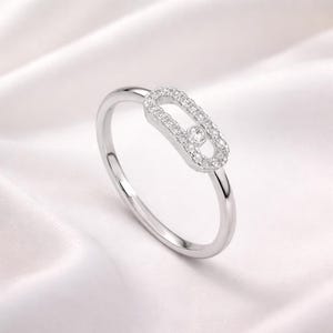 Puede incluir: Un anillo de plata con un dise&ntilde;o &uacute;nico. El anillo presenta un elemento central de forma ovalada incrustado con peque&ntilde;as piedras preciosas transparentes. Una sola gema m&aacute;s grande est&aacute; engastada en el centro del &oacute;valo. El anillo se encuentra sobre un fondo blanco y sedoso.