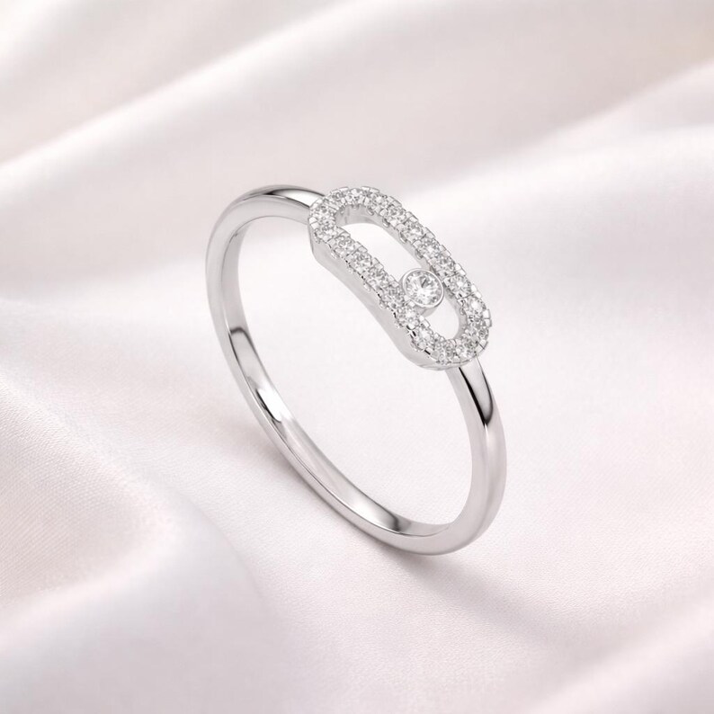 Puede incluir: Un anillo de plata con un dise&ntilde;o &uacute;nico. El anillo presenta un elemento central de forma ovalada incrustado con peque&ntilde;as piedras preciosas transparentes. Una sola gema m&aacute;s grande est&aacute; engastada en el centro del &oacute;valo. El anillo se encuentra sobre un fondo blanco y sedoso.