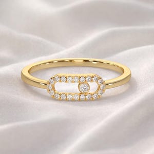 Puede incluir: Un anillo de oro con un dise&ntilde;o central rectangular. El centro est&aacute; incrustado con peque&ntilde;as gemas claras que rodean una gema individual m&aacute;s grande. La banda del anillo es delgada y pulida, sobre un fondo blanco y sedoso.