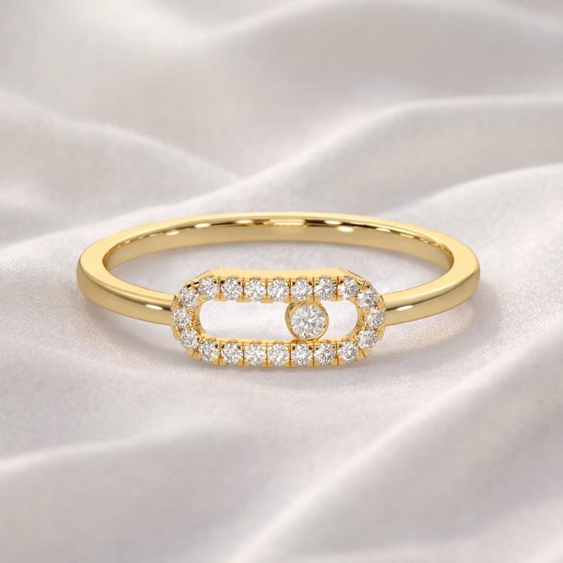 Puede incluir: Un anillo de oro con un dise&ntilde;o central rectangular. El centro est&aacute; incrustado con peque&ntilde;as gemas claras que rodean una gema individual m&aacute;s grande. La banda del anillo es delgada y pulida, sobre un fondo blanco y sedoso.