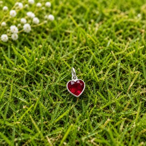 May include: A silver pendant featuring a faceted, heart-shaped red gemstone. The pendant has a small, circular silver bail at the top for attachment. The piece is positioned centrally, showcasing the vibrant red colour and the metallic silver setting.