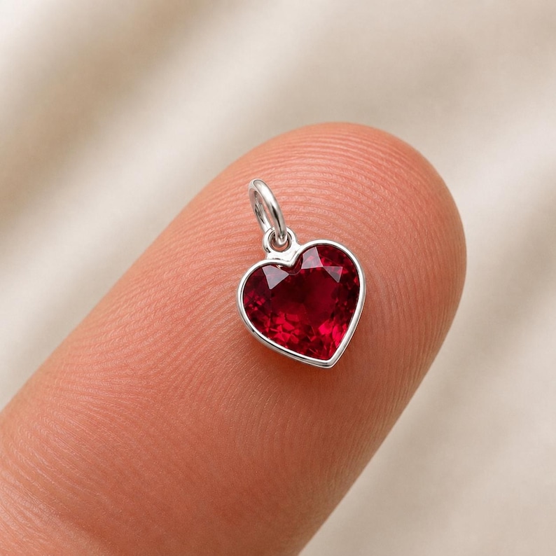 May include: A small heart-shaped pendant featuring a deep red faceted gemstone set in a polished silver-toned metal frame. The charm includes a small circular jump ring at the top for attachment, showcasing a clear, vibrant red colour and precise cut.