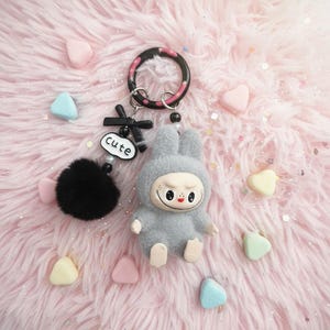May include: A gray bunny keychain with a smiling face, black pom-pom, and the word "cute" on a white oval. The keychain has a black and pink ring and is surrounded by heart-shaped candies on a pink, fluffy surface.