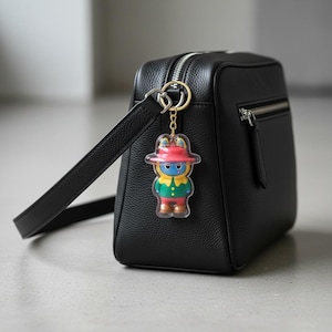 May include: A black leather handbag with a gold zipper and a small, colorful keychain. The keychain features a cartoon character in a red hat, green jacket, and blue face. The bag has a shoulder strap.