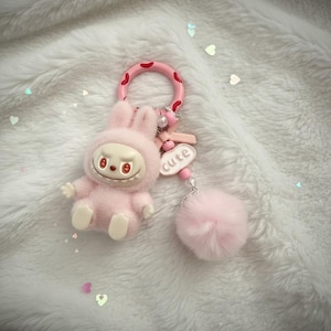 May include: A pink keychain featuring a plush bunny figure with a smiling face and a fluffy pom-pom. The keychain has a pink ring with red spots and a charm that says "cute".