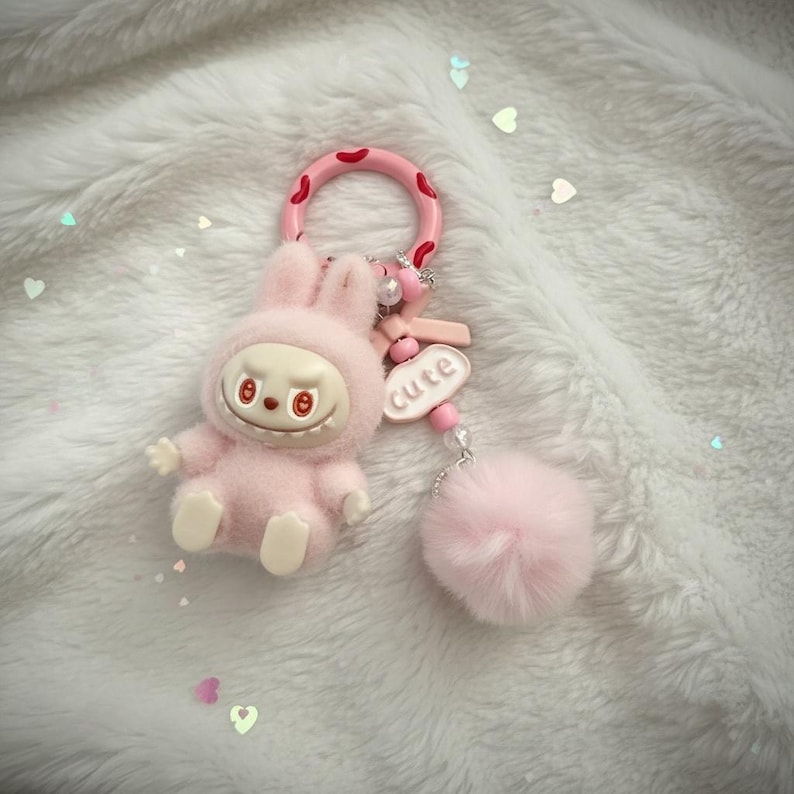 May include: A pink keychain featuring a plush bunny figure with a smiling face and a fluffy pom-pom. The keychain has a pink ring with red spots and a charm that says "cute".