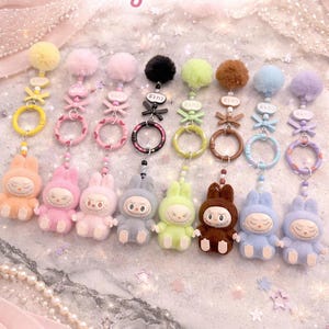 May include: A collection of plush keychains in various colors, including peach, pink, gray, and brown. Each keychain features a bunny-like character with a pom-pom and decorative accents. The text "Cute Plush Keychains" is visible.