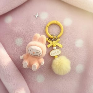 May include: A peach-colored bunny figurine with closed eyes and a matching hooded outfit. A yellow keychain with a bow, the word "cute", and a fluffy pom-pom is next to the bunny. The background is a soft pink.