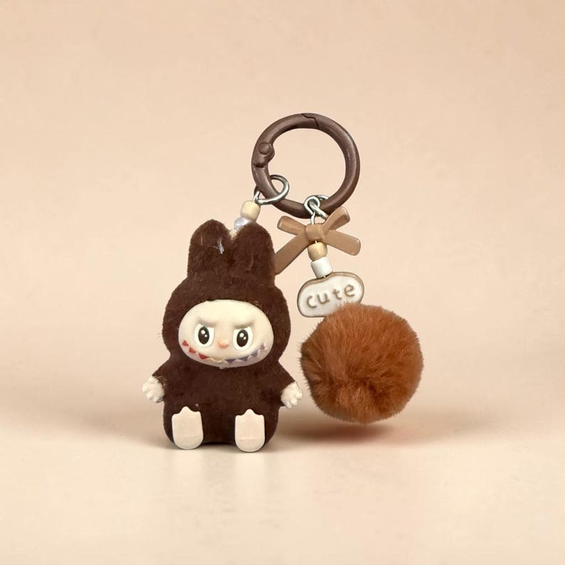 May include: A brown bunny keychain with a pom-pom and a metal ring. The bunny is wearing a hooded outfit with a white face and a stitched mouth. A small tag with the word "cute" is attached.
