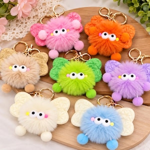 May include: A collection of seven plush pom-pom keychains, each featuring a fluffy round body, two large white eyes, and two small pom-pom feet. Each keychain has a pair of patterned wings attached to the back and a gold-tone metal keyring.