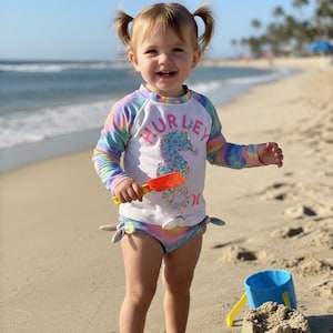May include: A young child wearing a white and rainbow-colored rash guard and matching swim bottoms with the word "HURLEY" and a seahorse graphic. The child is on a sandy beach, holding a small orange shovel.