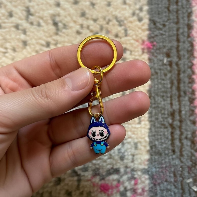 May include: A gold-colored keychain with a cartoon character charm. The charm is a blue figure with a dark blue head, white eyes, and a toothy grin. The keychain has a gold ring and clasp.